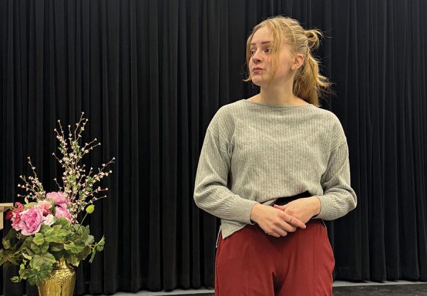 Photo of a play scene depicting a young woman looking past flowers