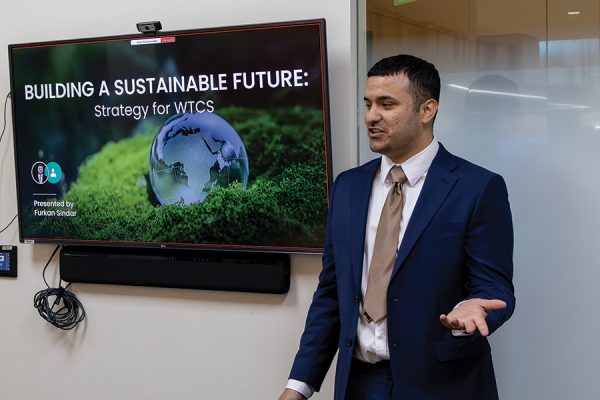 A young man in a blue blazer speaks in front of a TV monitor with the phrase, "Building a Sustainable Future," on the screen.