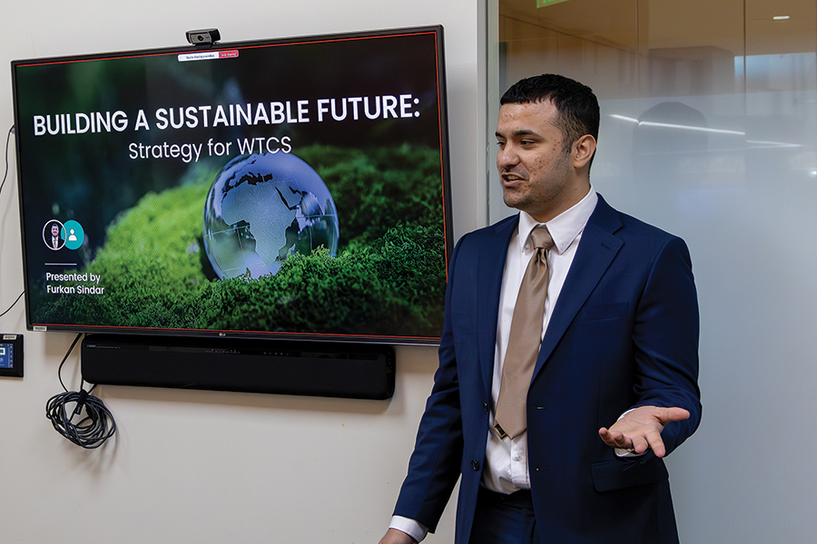 A young man in a blue blazer speaks in front of a TV monitor with the phrase, "Building a Sustainable Future," on the screen.