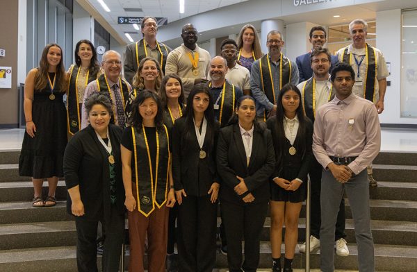 A group of staff, faculty and students pose on a stairwell for a photograph wearing golden and black stoles.