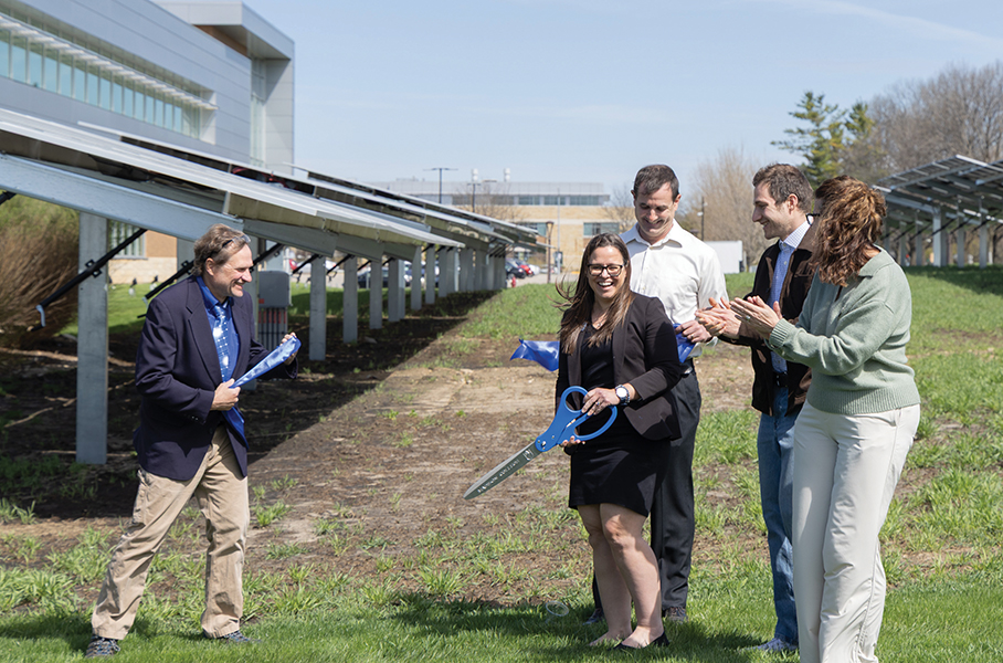 Five people smile and clap after cutting a ribbon between two rows of free-standing solar panels outside a Madison College building.