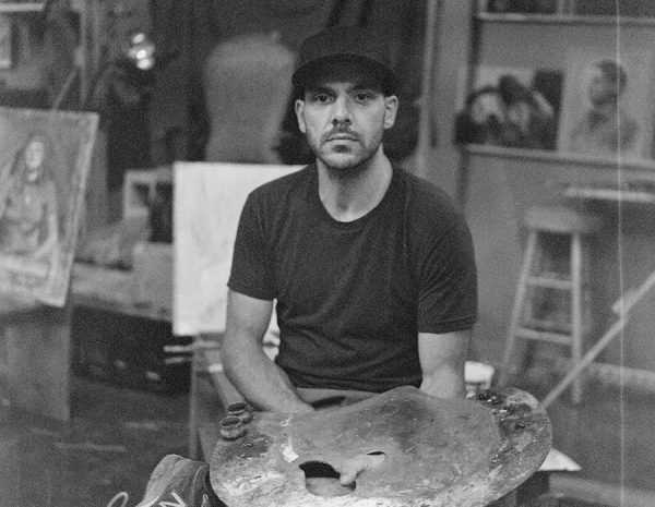 An artist holds an easel while sitting on a stool surrounded by paintings.
